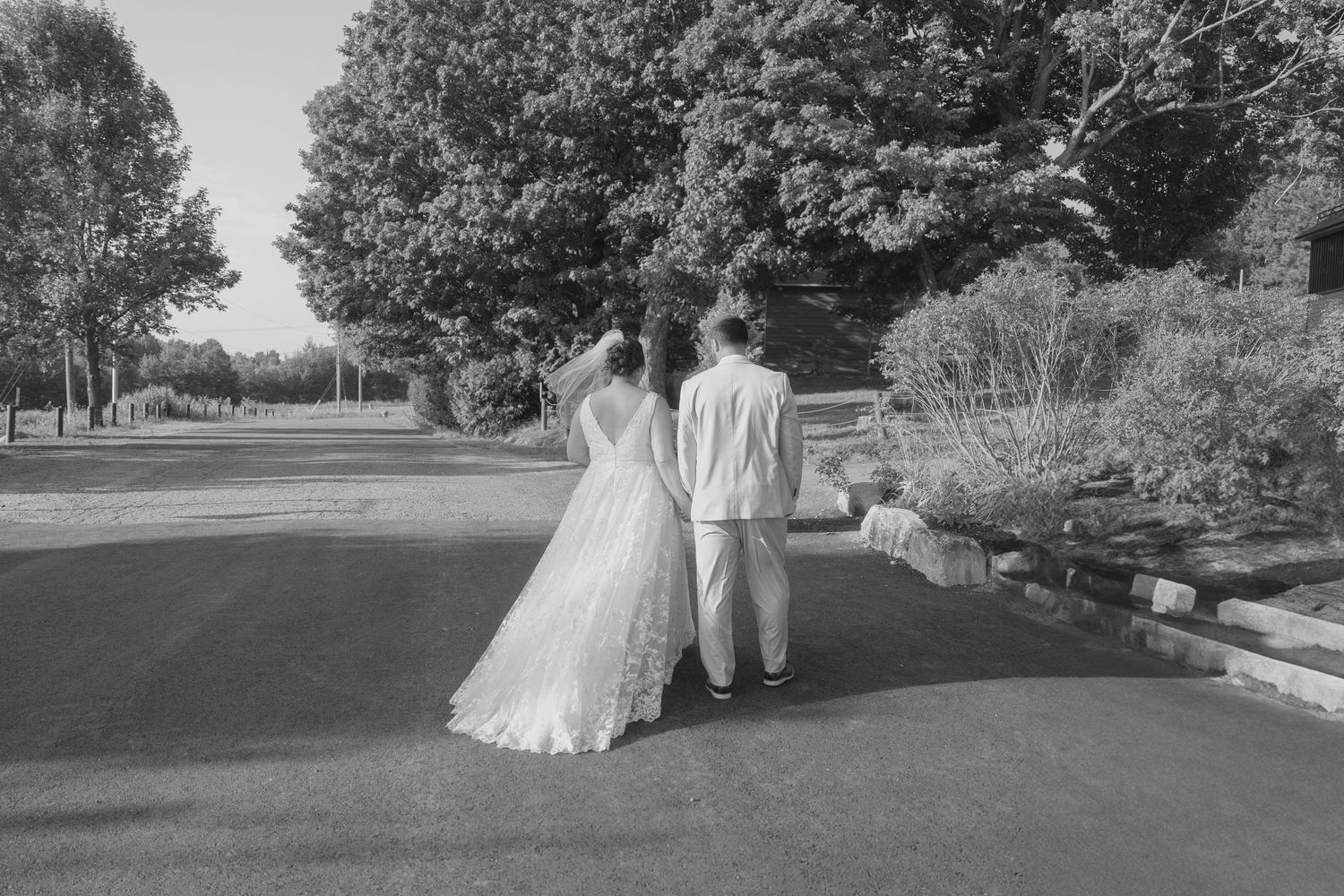 A couple walks hand in hand down a tree-lined path in black and white, with the bride wearing a flowing white dress.