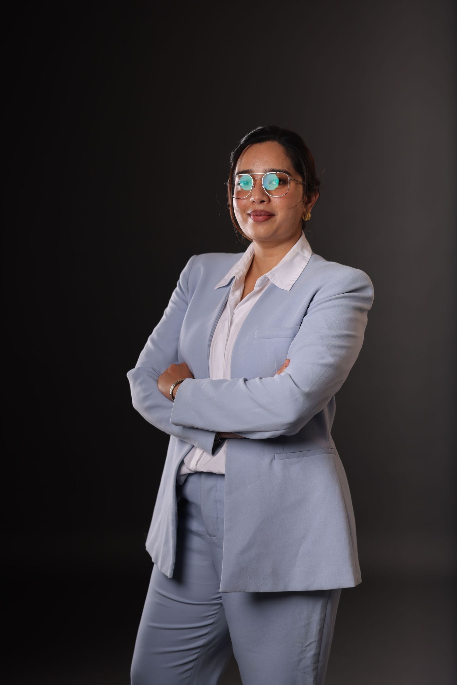 Professional woman in light blue suit with crossed arms and green glasses posing confidently against dark background.