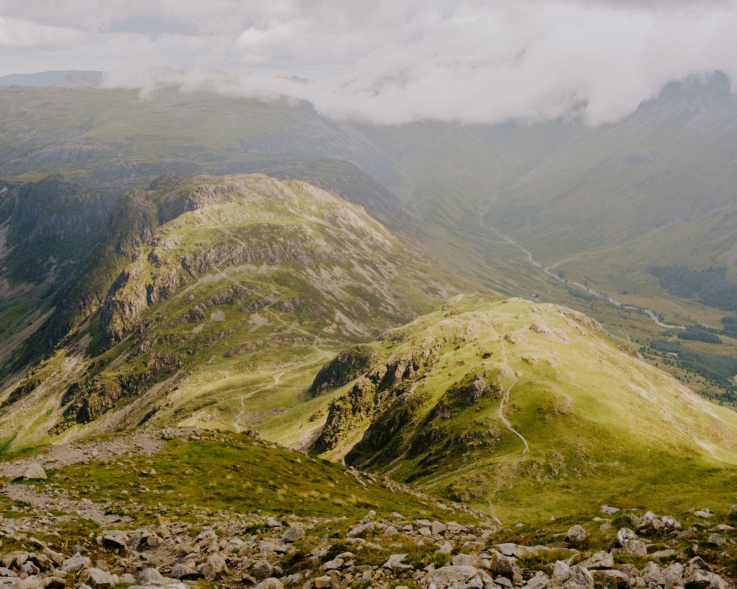 Aerial view of lush green mountain valleys and rocky peaks under a cloudy sky in a scenic highland landscape.