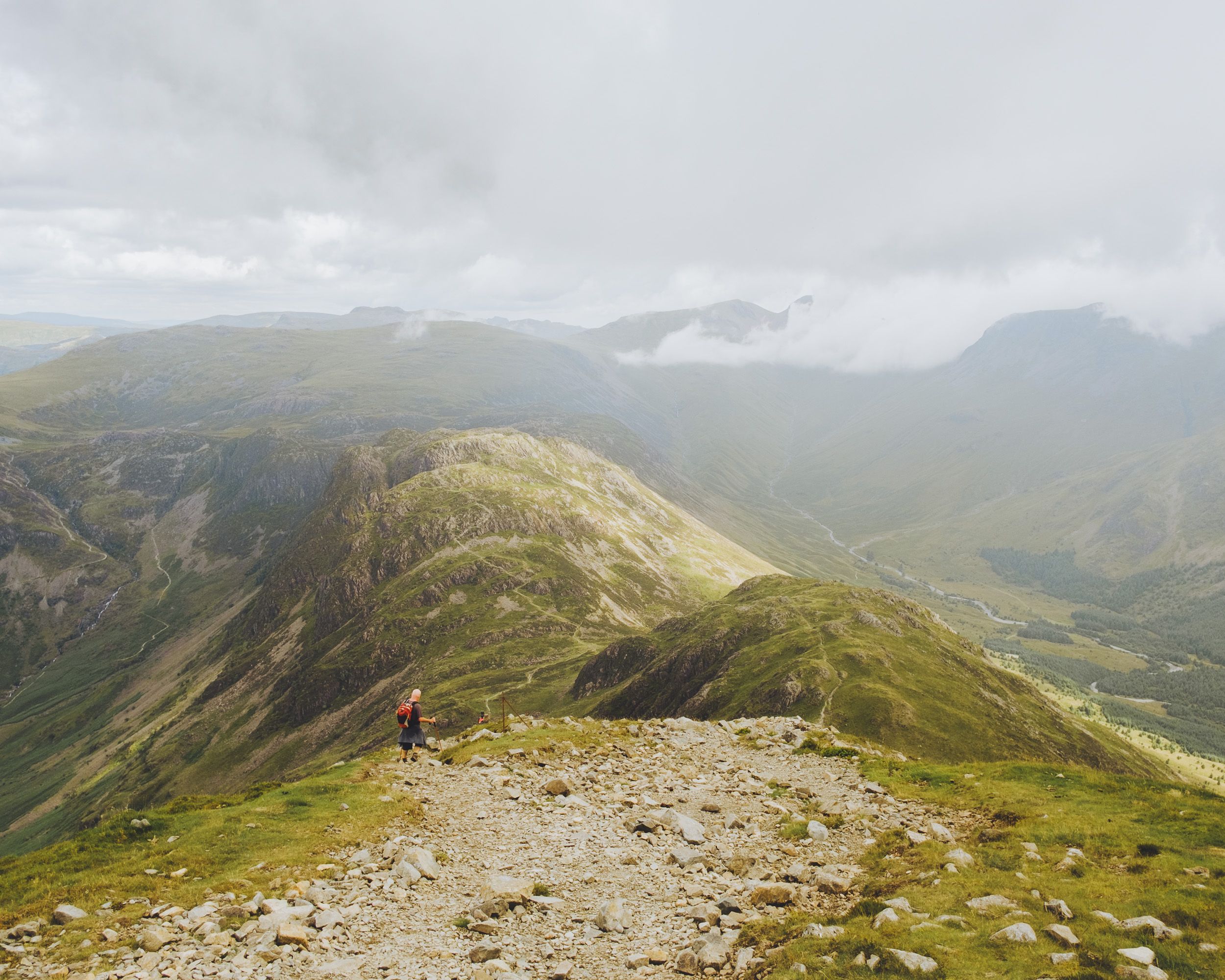Hiker on a rocky mountain ridge with sweeping green valley views under cloudy skies.