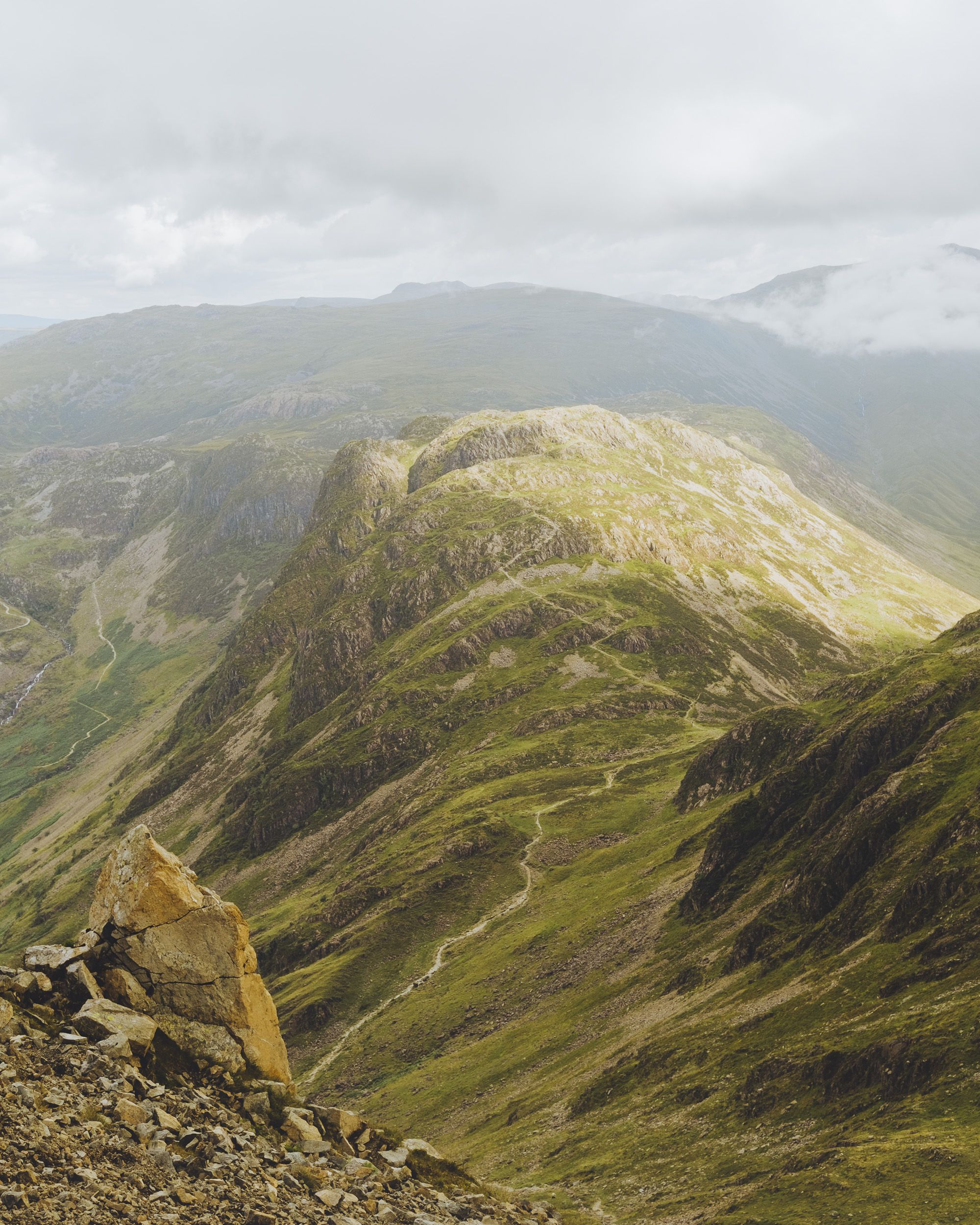 Dramatic green mountain landscape with rocky peaks, winding paths, and low clouds over misty highlands.