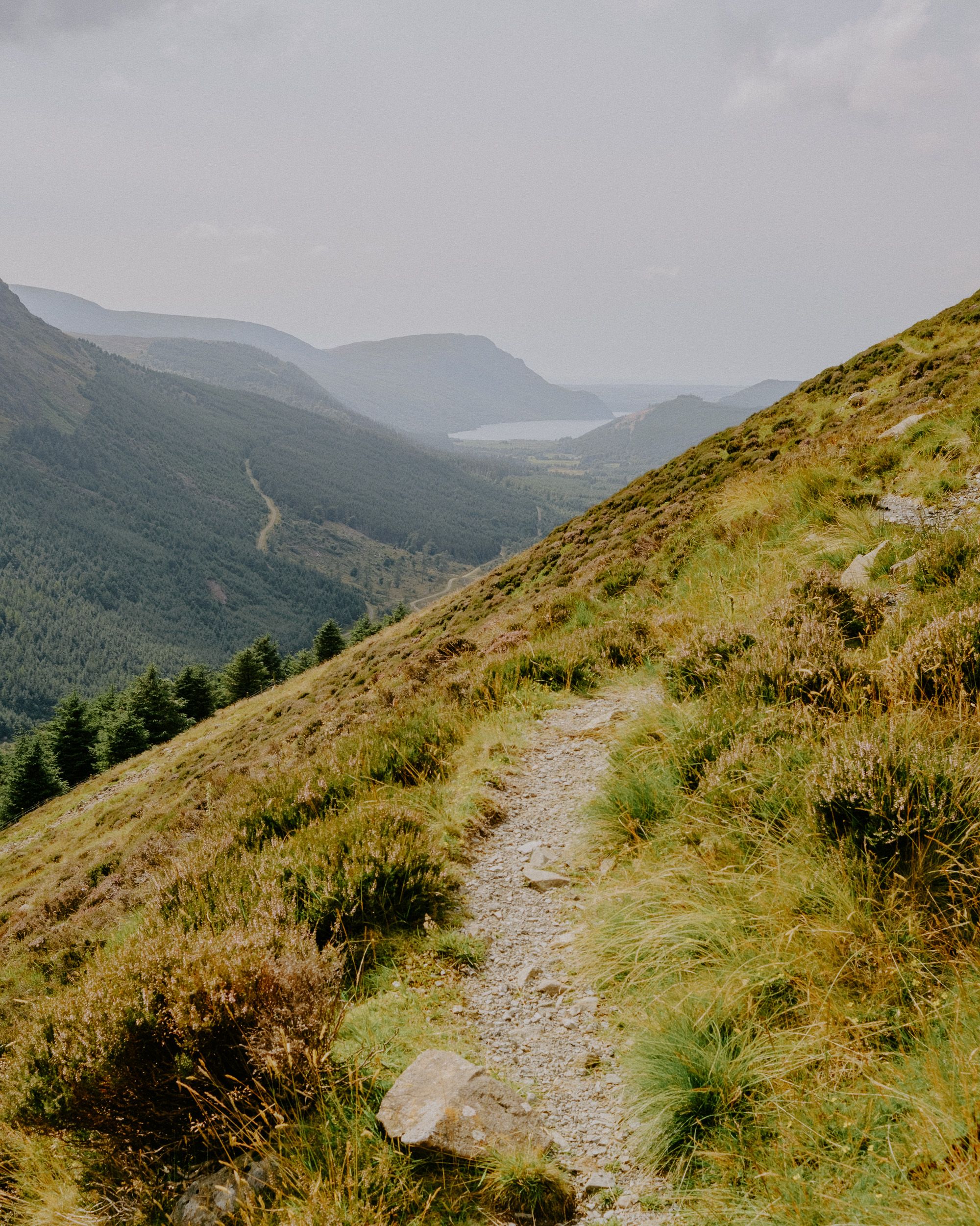 A narrow gravel hiking trail winds along a lush green mountain hillside with a scenic valley and lake in the distance.
