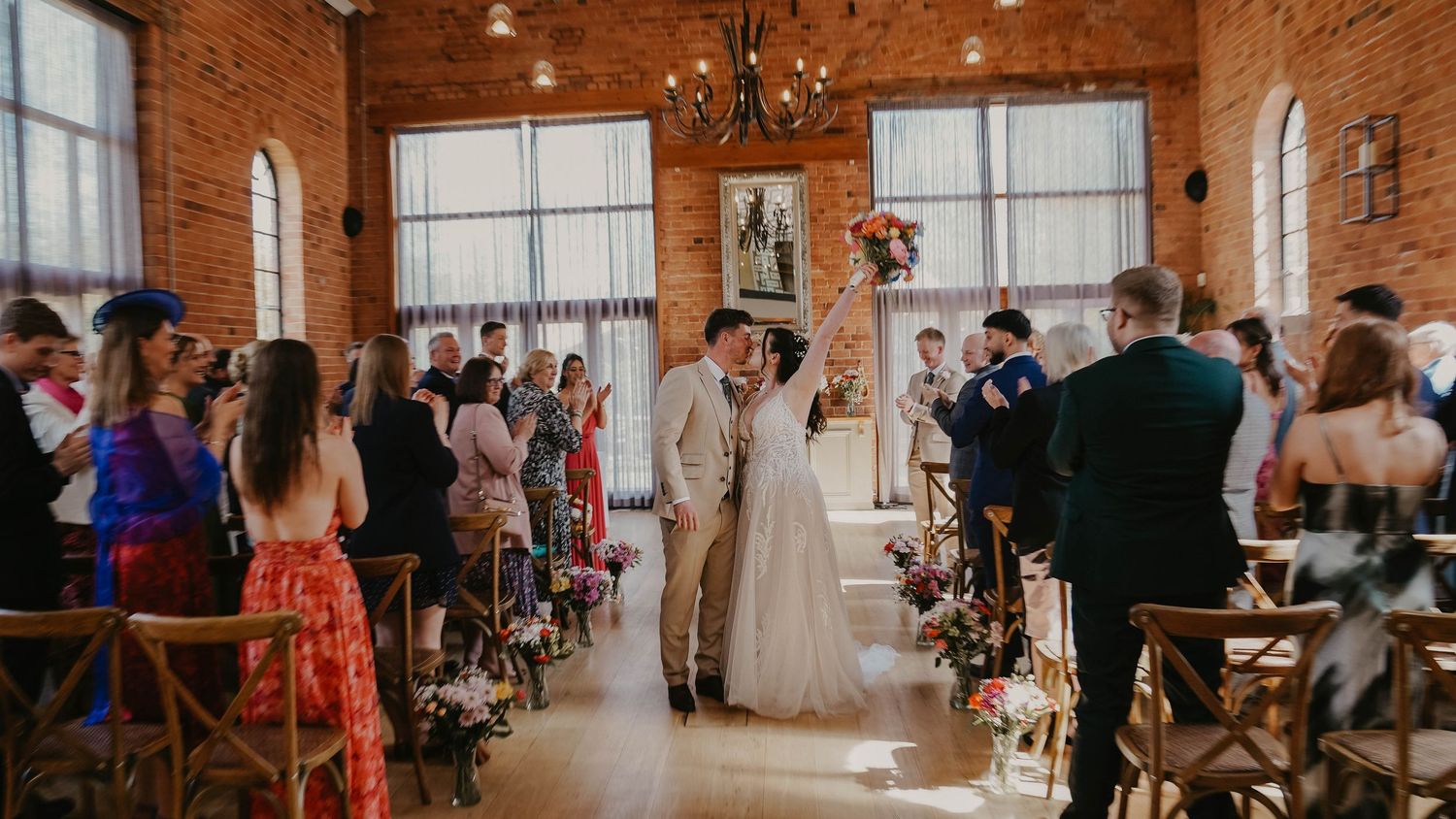 Couple shares first kiss at rustic brick wedding venue with floral aisle decorations and guests watching.