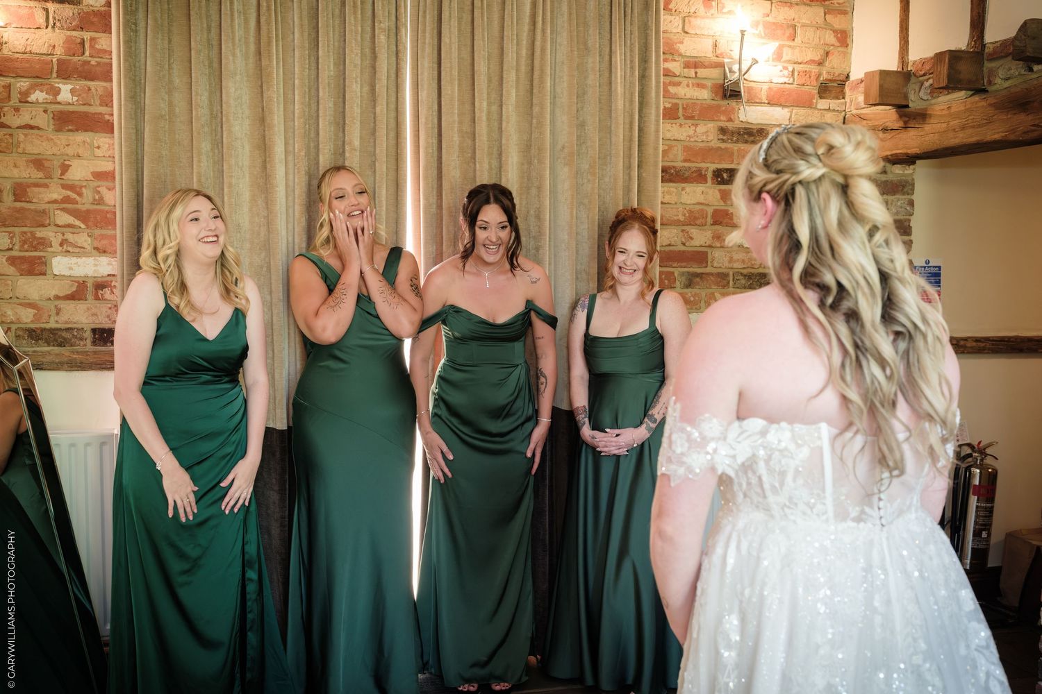 Bridesmaids in emerald green gowns react with joy and surprise as bride in white lace dress reveals her look before wedding.