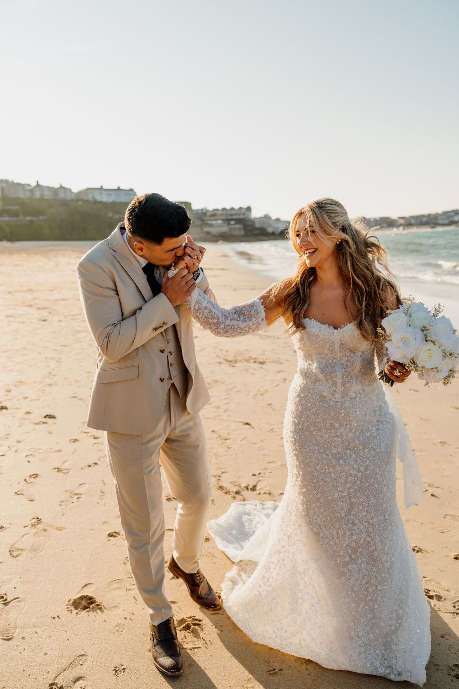 Groom in beige suit kisses bride's hand on sunny beach, bride smiles in lace wedding gown holding white bouquet.