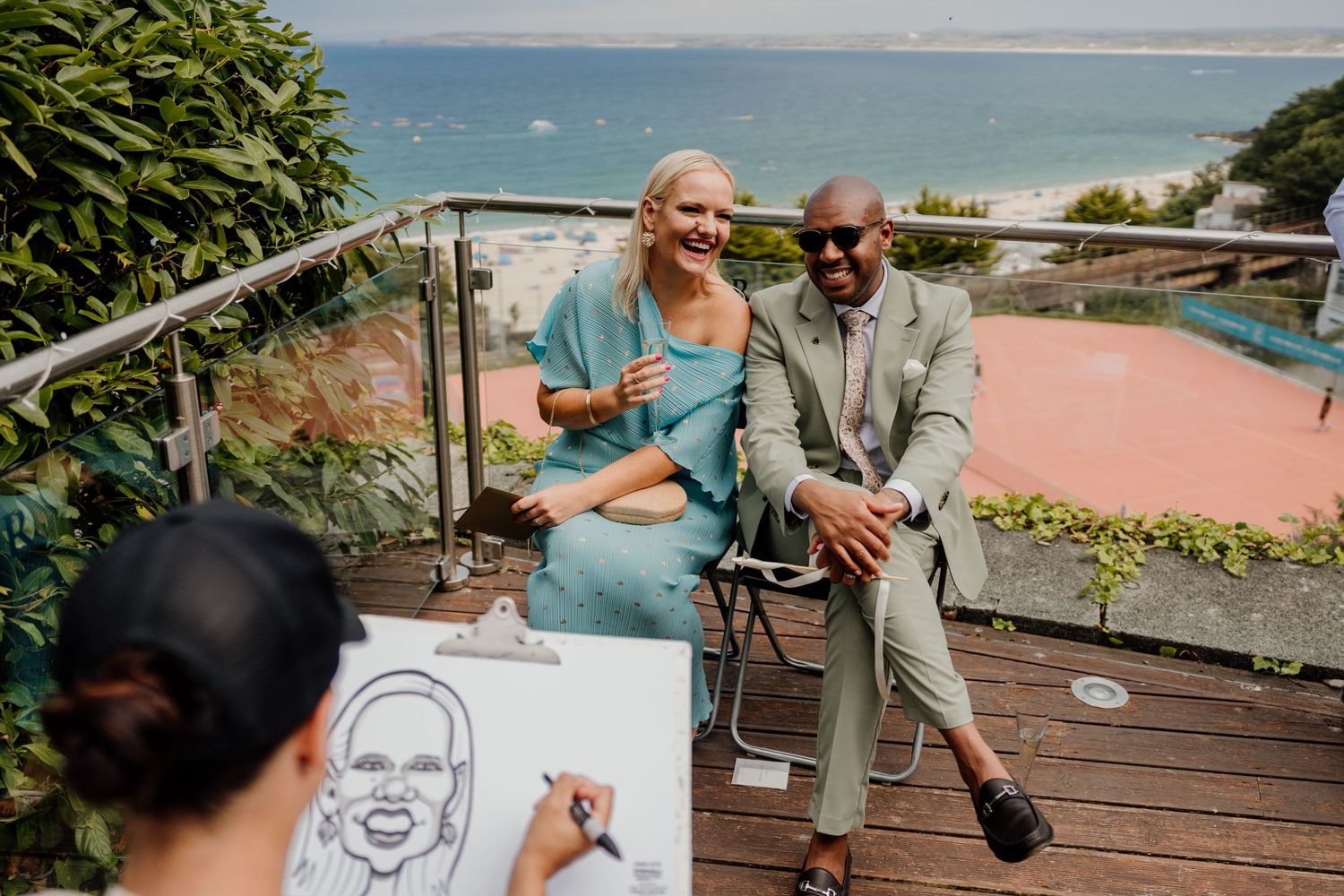 Couple laughing as caricature artist sketches their portrait on a scenic coastal terrace at an outdoor event.