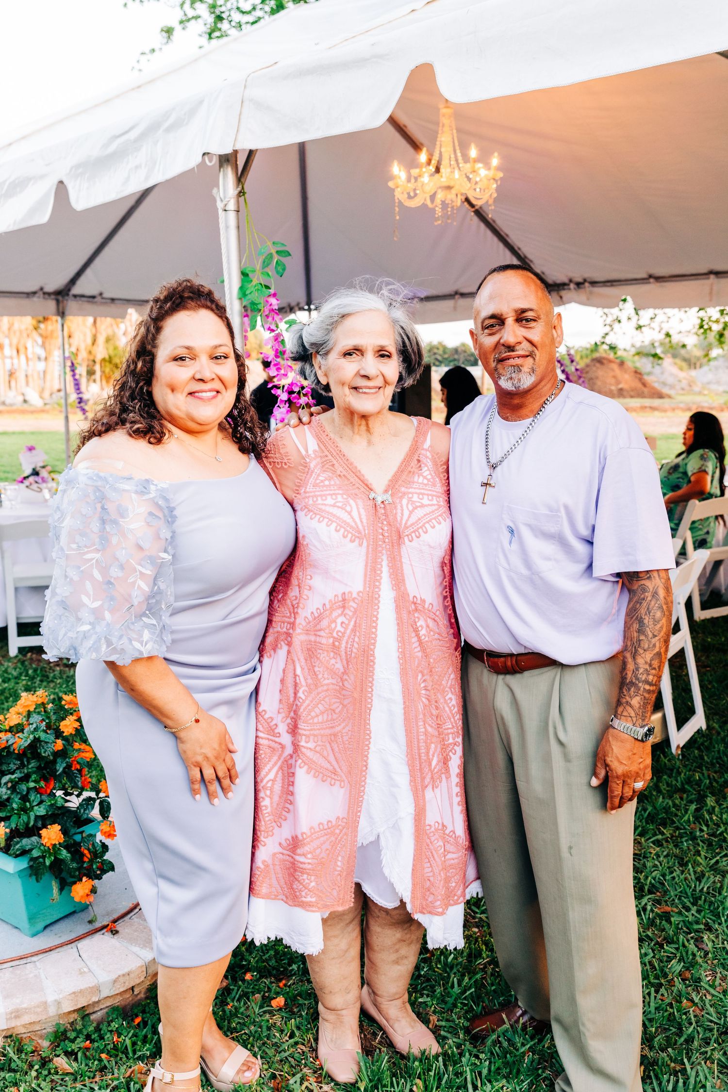 Three people smiling together under a white tent at an outdoor event, dressed in elegant attire for a celebration.