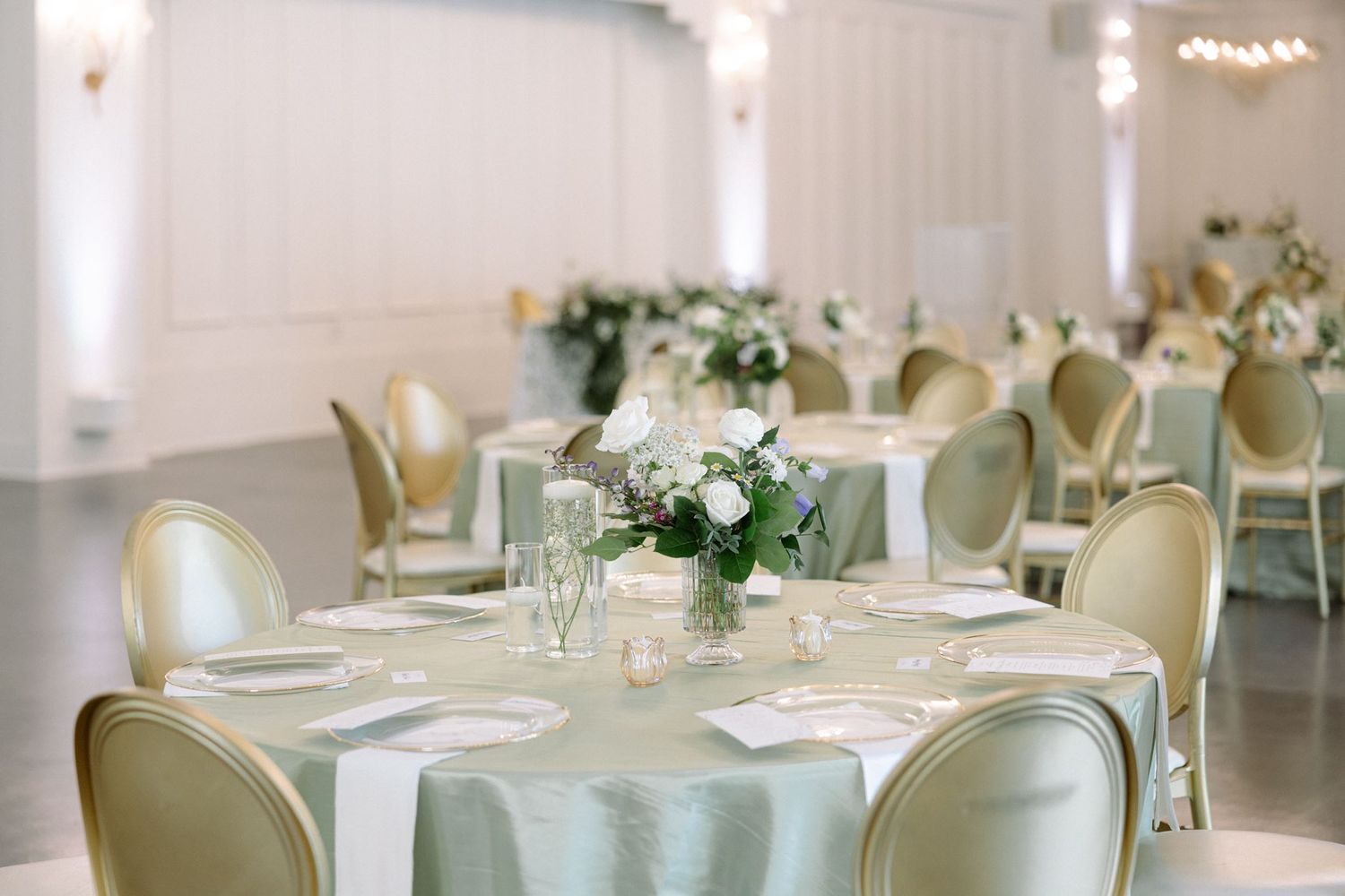Elegant wedding reception hall with sage green tablecloths, gold chairs, and white floral centerpieces in a bright ballroom.