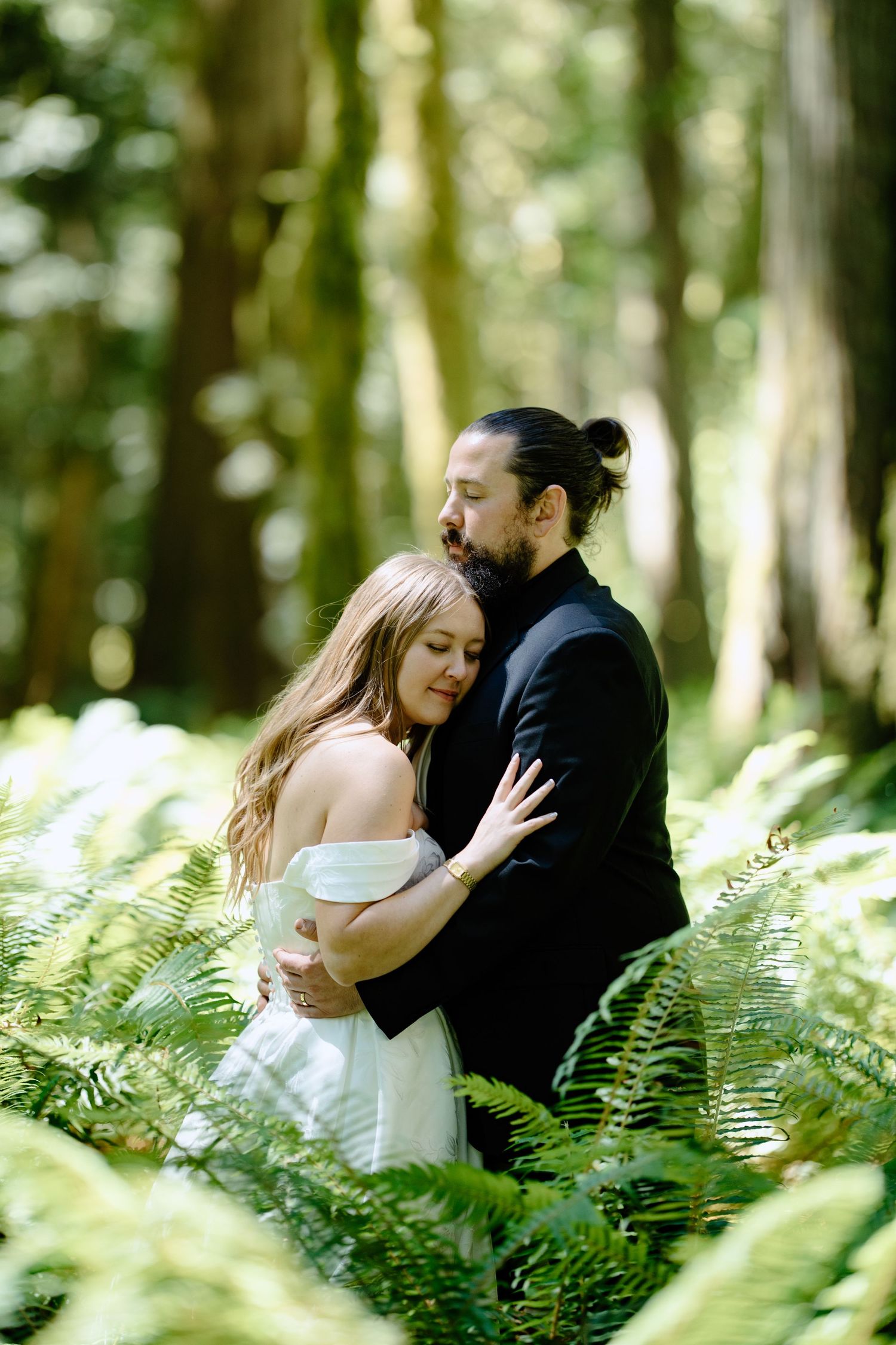 Couple embracing in a lush forest surrounded by ferns, bride in white off-shoulder dress, groom in black suit.