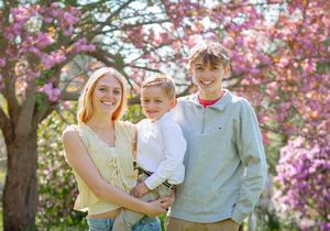 Two teenagers and a young child pose together outdoors among blooming pink cherry blossom trees in spring.