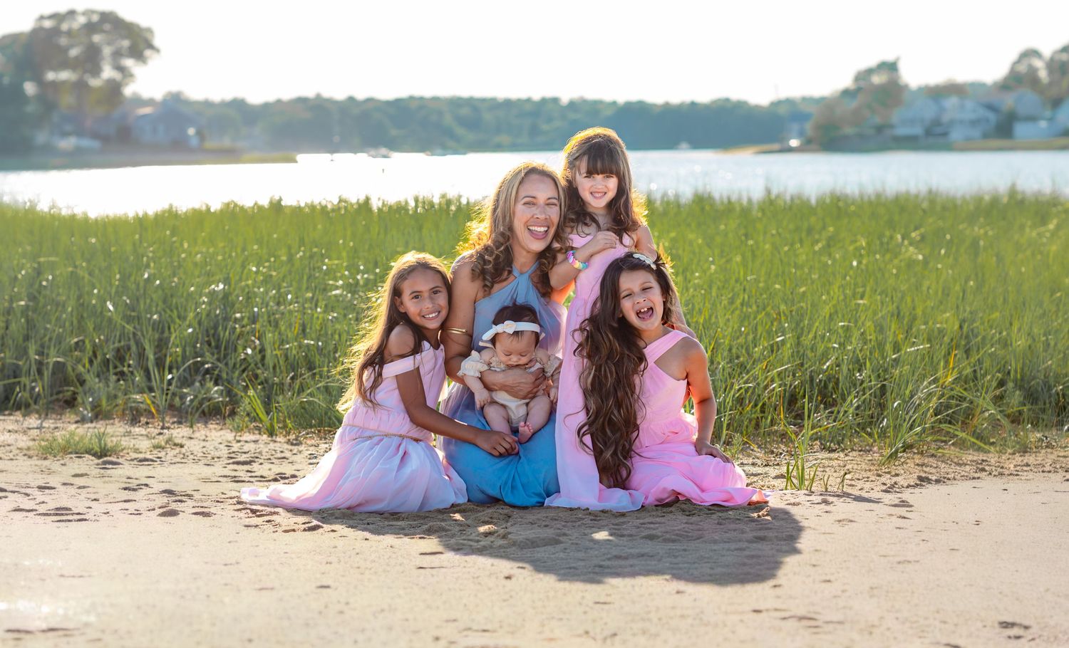 A woman and four girls in colorful dresses pose together on a sandy beach near a grassy waterfront at sunset.