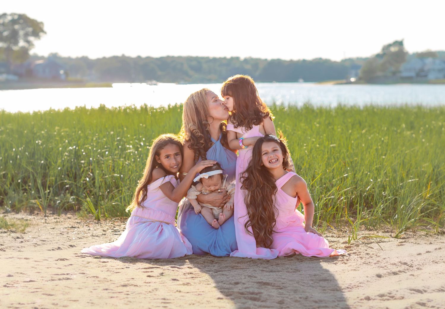 A mother sits on a sandy beach with her four daughters in pink dresses, sharing a joyful moment near a sunlit river.