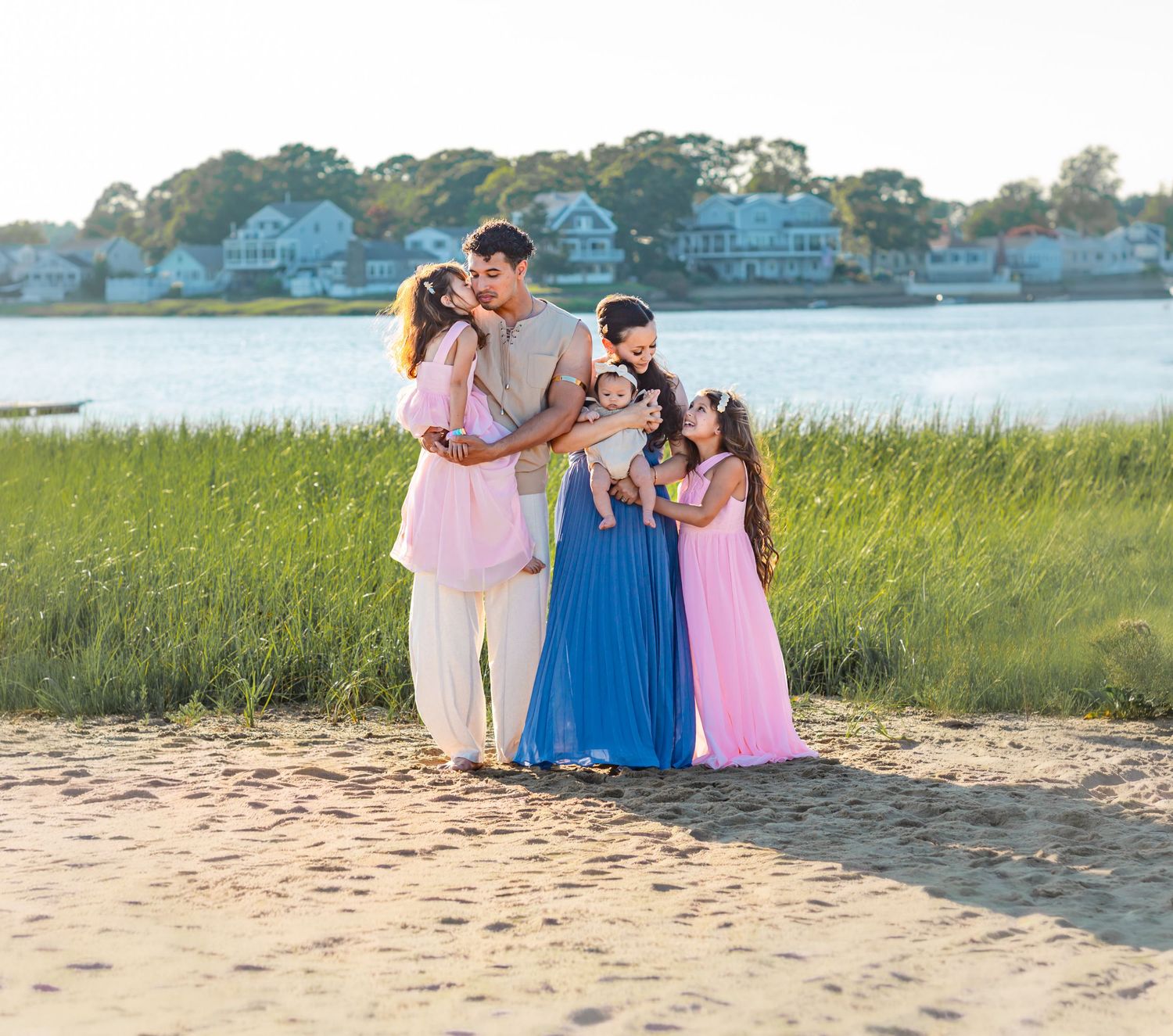 Family of five posing on a sandy beach at golden hour, with tall grass and waterfront homes in the background.
