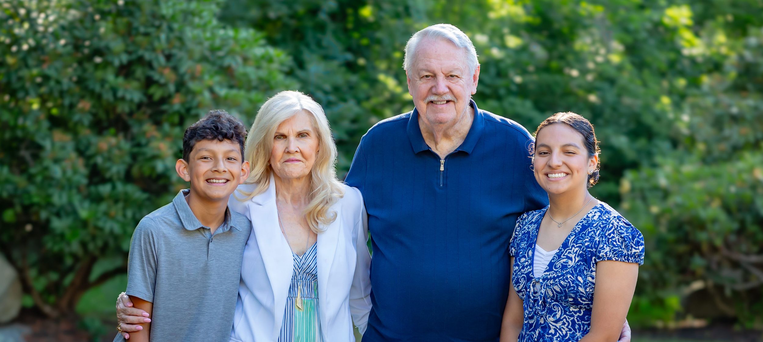 Two grandparents smiling outdoors with two grandchildren, surrounded by lush green trees in a family portrait.