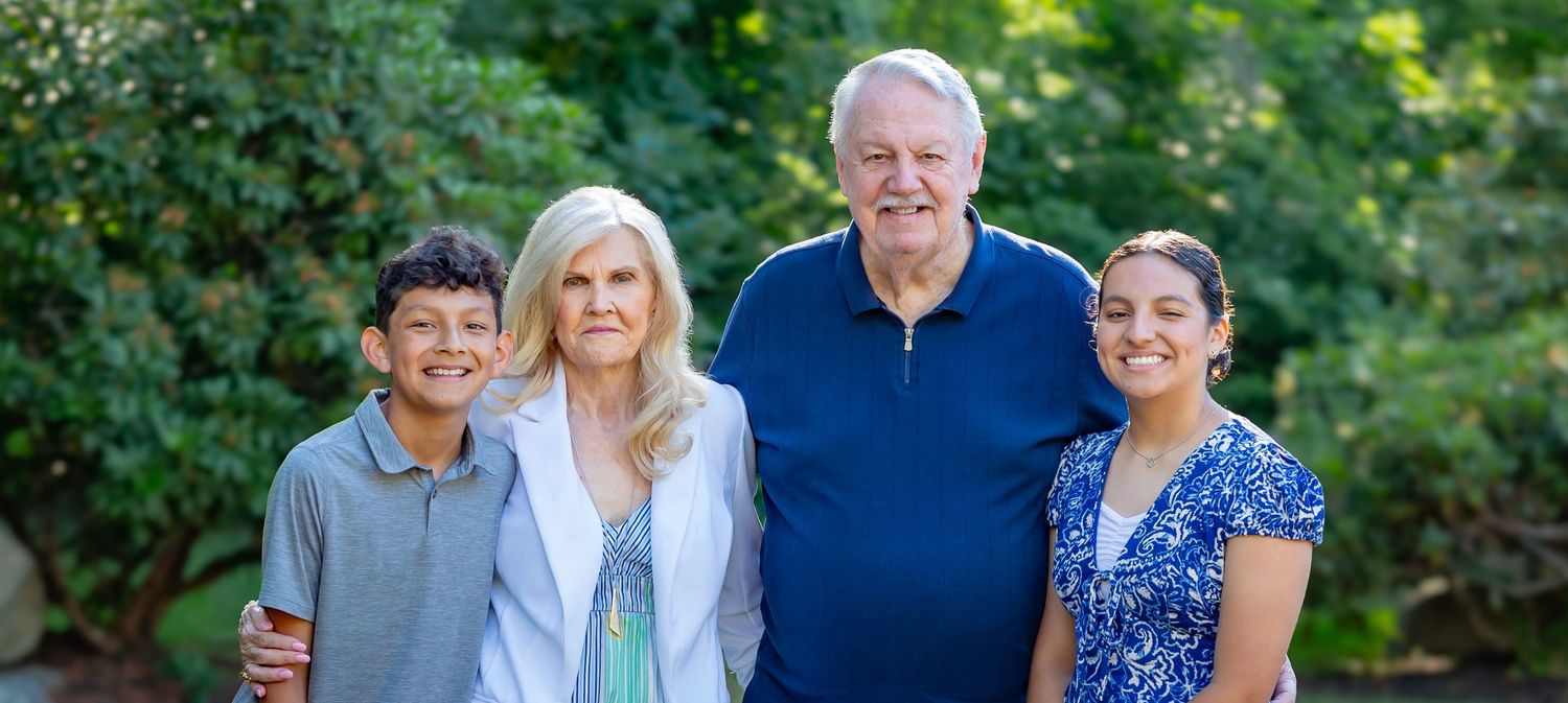 Two grandparents smiling outdoors with two grandchildren, surrounded by lush green trees in a family portrait.