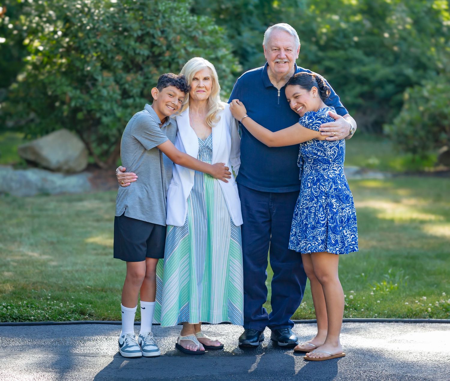 Family of four smiling and hugging outdoors, with lush green trees in the background on a sunny day.