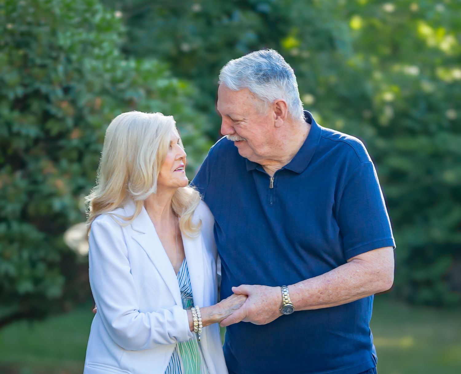 Elderly couple smiling at each other outdoors, woman in white jacket and man in navy polo, surrounded by green trees.