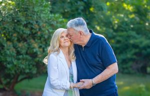 An older couple sharing a tender kiss outdoors, surrounded by lush green trees in a garden setting.
