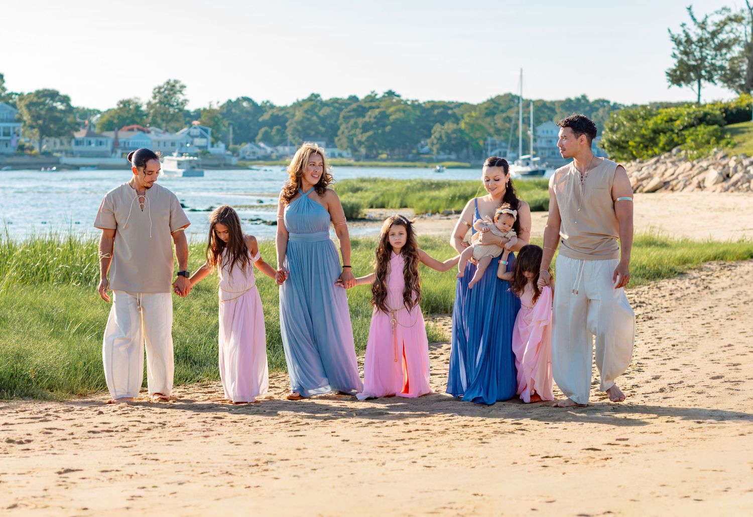 Family of seven posing on sandy beach near water, wearing coordinated pastel outfits in pink, blue, and beige tones.