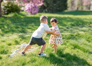 Two young children playing together on green grass in a sunny park with blooming pink trees in the background.
