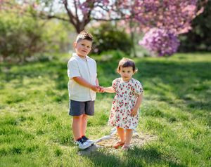 Two young children holding hands on green grass with pink flowering trees in a sunny garden setting.