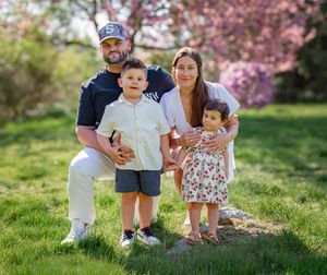 Family of four posing outdoors in a blooming spring park, parents with young son and toddler daughter smiling.
