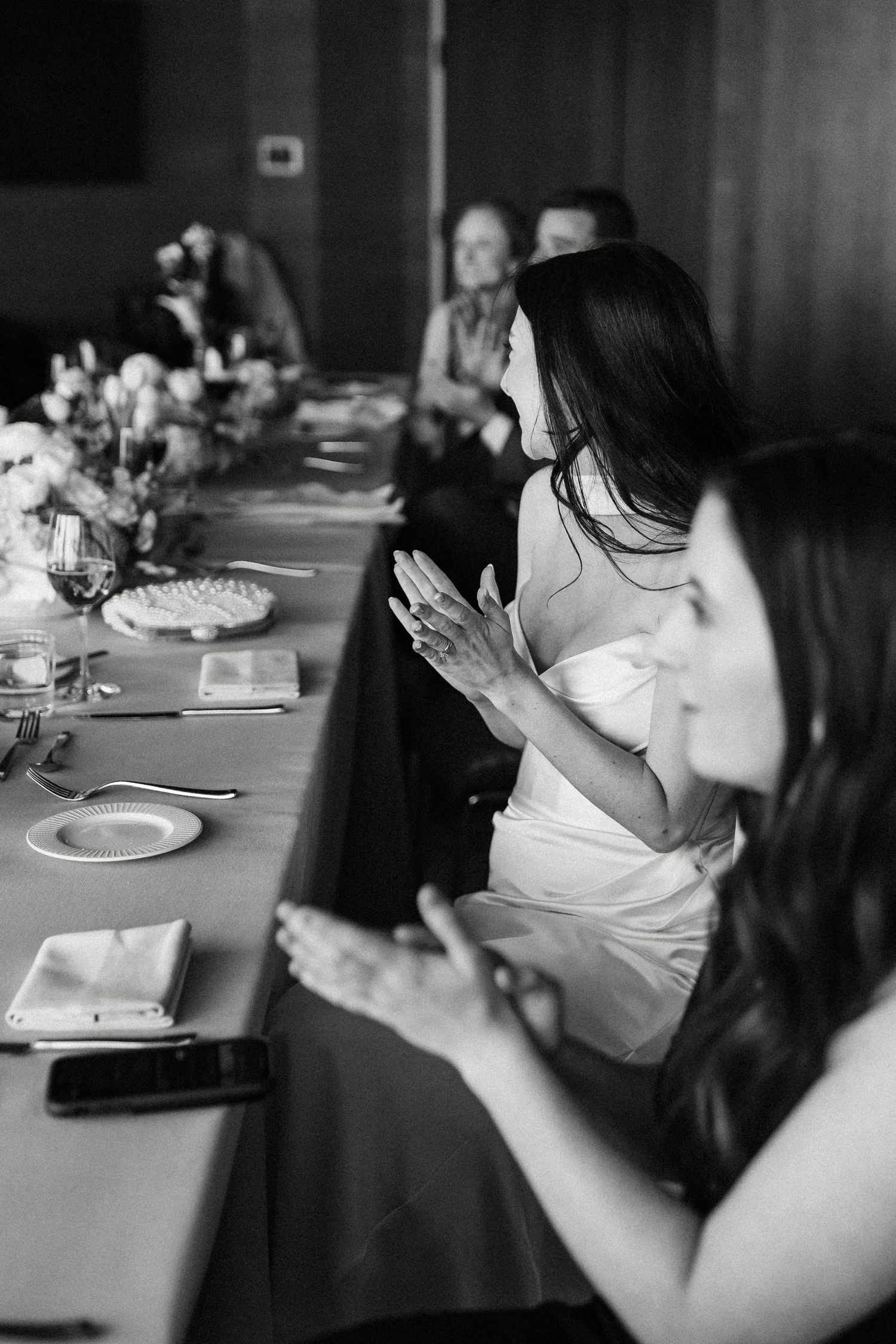 Bride and groom seated at wedding reception head table, smiling during toasts in elegant venue.