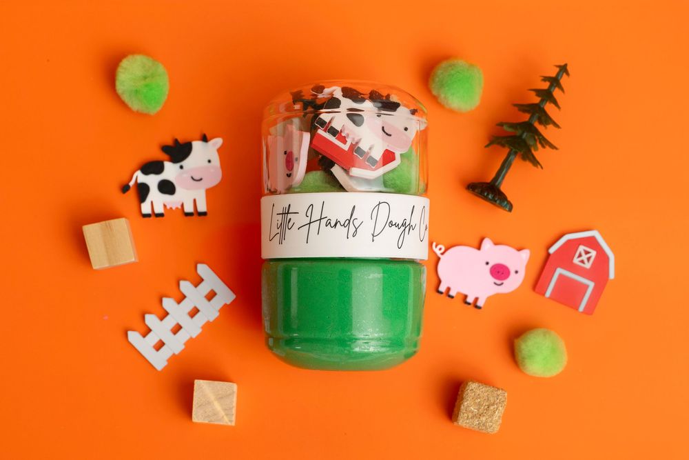 Little Hands Dough kit in green container with farm-themed accessories including cow, pig, barn, fence, and tree on orange background.