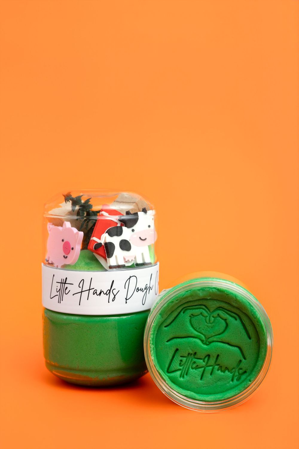 Little Hands Dough green play dough jar with farm animal toppers including pig and cow on orange background.