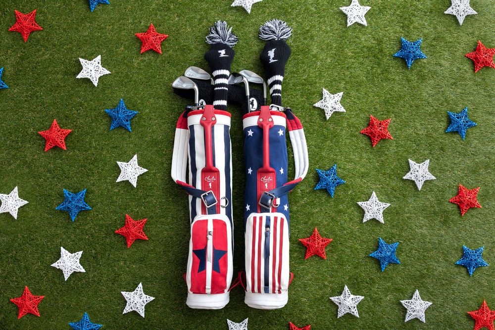 Two patriotic red, white, and blue golf bags with club covers on green turf surrounded by star-shaped decorations.