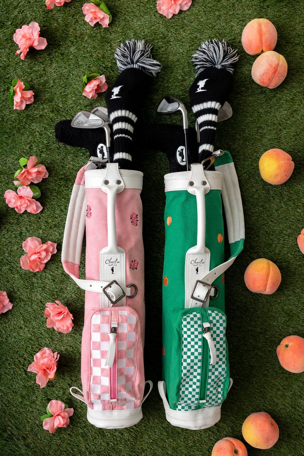 Two stylish golf bags in pink and green with checkered pockets, black-striped headcovers, surrounded by peaches and pink flowers on grass.