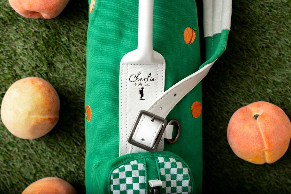 Green peach-print personalized golf bag with white leather strap, checkered pocket, and Charlie name tag on grass.