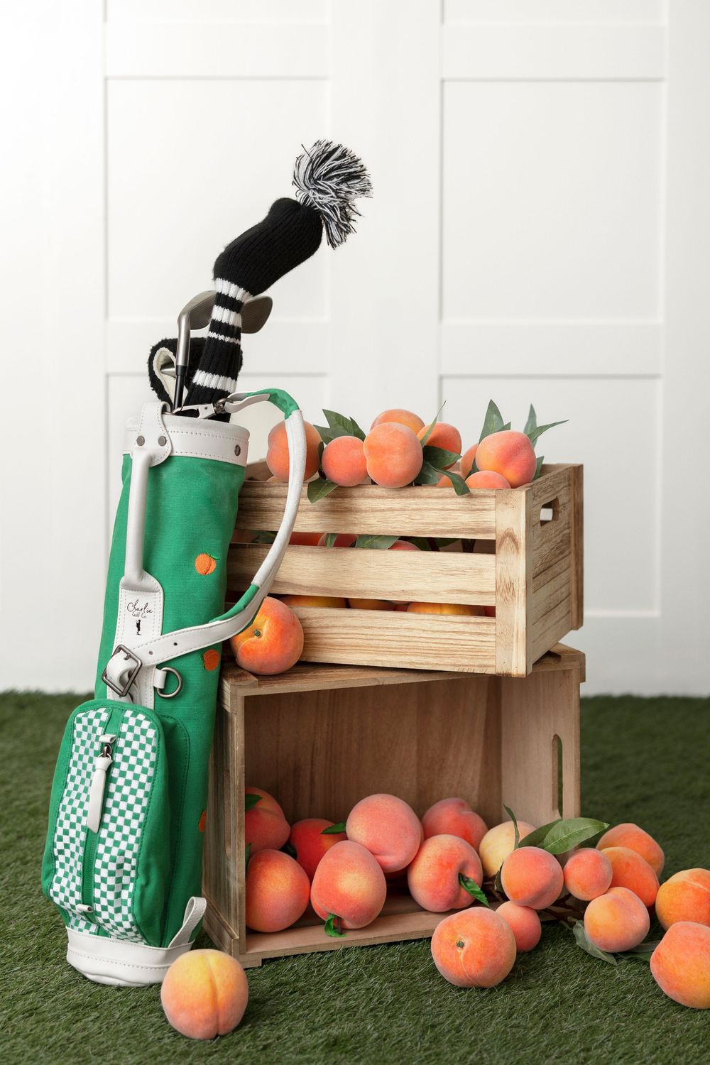 Green golf bag with clubs leaning against wooden crates filled with fresh peaches on artificial grass.