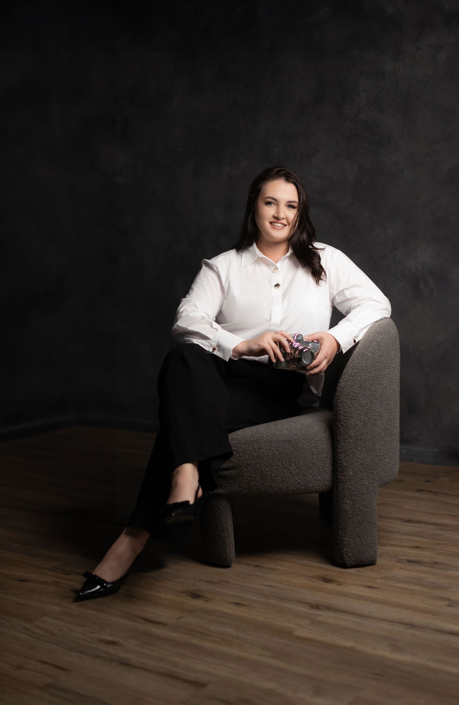 Professional portrait of person in white blouse sitting relaxed in gray armchair against dark background.