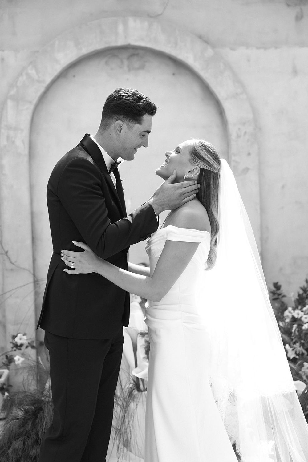 Black and white photo of a groom in a tuxedo tenderly holding his bride's face as she gazes up at him in a wedding dress.