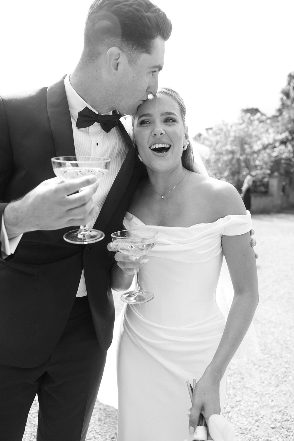 Black and white photo of a laughing bride in off-shoulder gown and groom in tuxedo toasting with champagne glasses.
