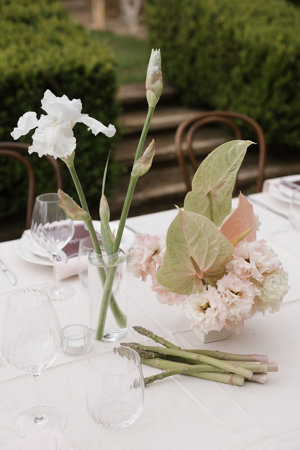 Elegant outdoor wedding table with white iris flowers, pink blooms, green anthurium leaves, and asparagus stems in glass vases.