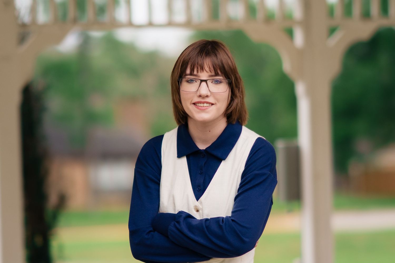 Young woman with glasses and brown hair poses arms crossed under a wooden pergola outdoors.