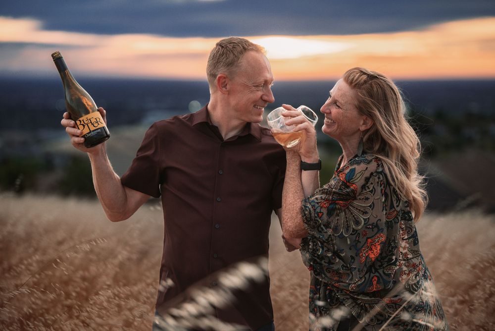 Couple celebrating at sunset in a field, man holding wine bottle, woman sipping from glass, smiling joyfully.