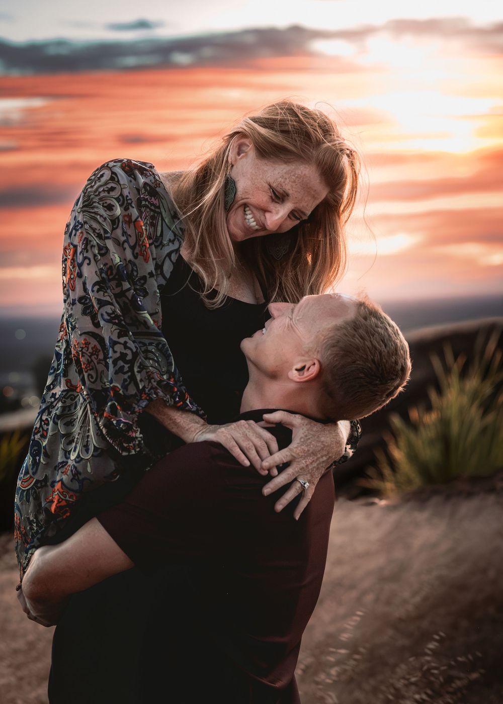 Couple sharing a joyful romantic moment at sunset, man lifting woman as she laughs against a vibrant orange sky.