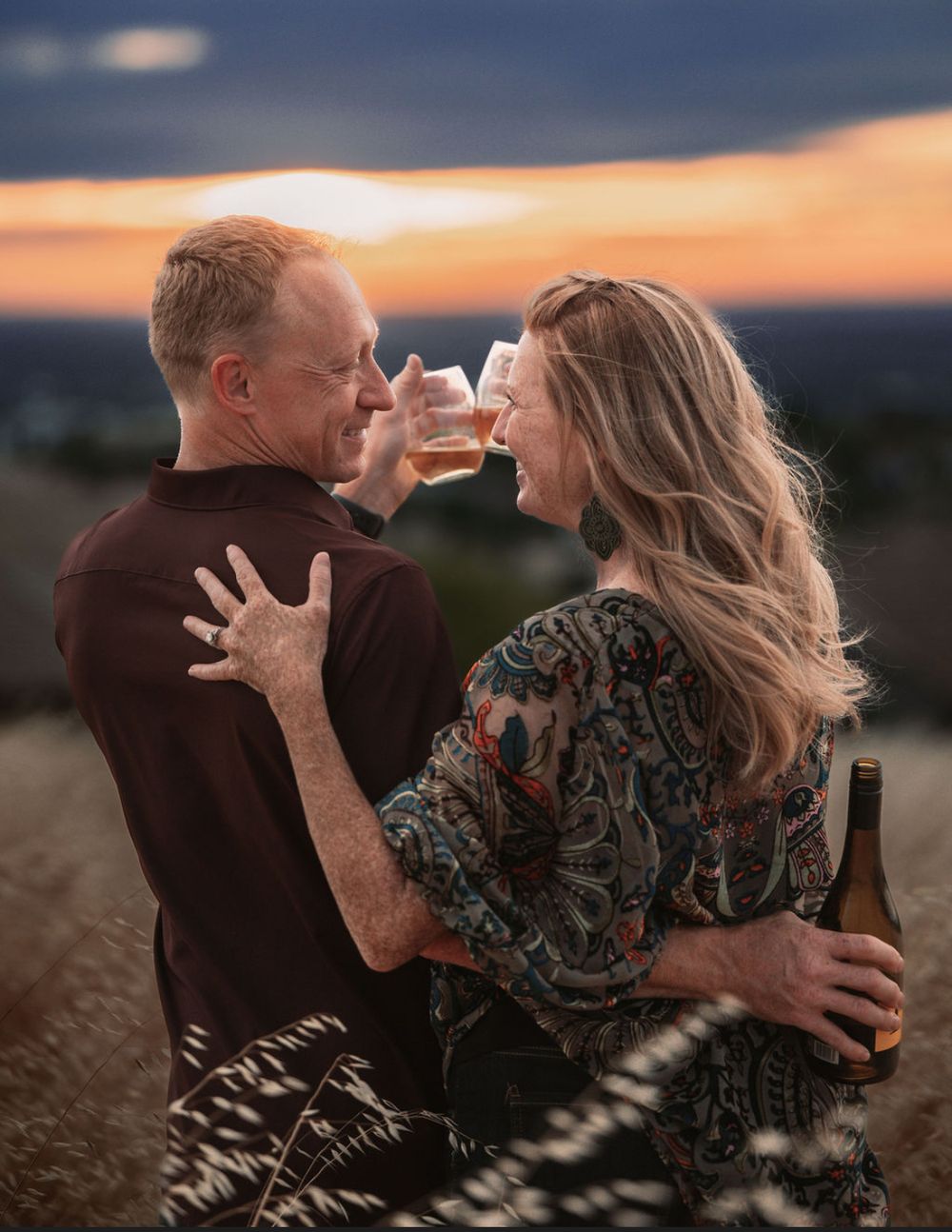 Couple sharing a romantic toast with beer glasses at sunset, embracing outdoors with a golden sky backdrop.