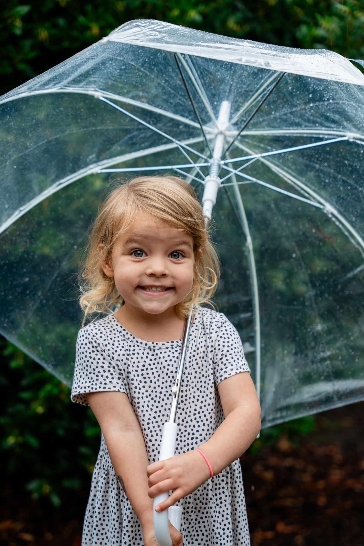 Smiling young blonde girl holding a clear transparent umbrella outdoors in the rain, wearing a white polka dot dress.