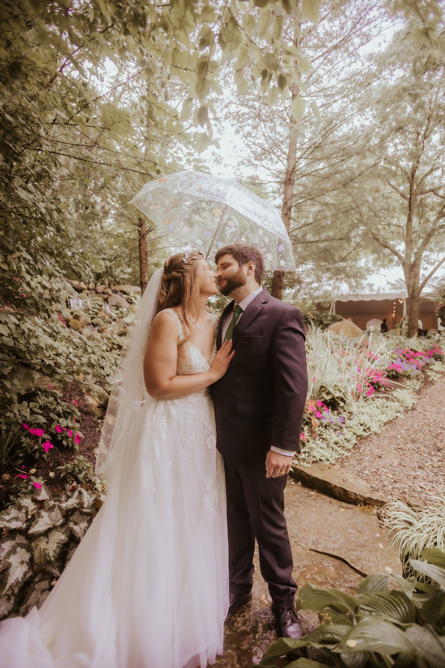 Bride and groom share a romantic moment under a clear umbrella in a lush garden setting on their wedding day.