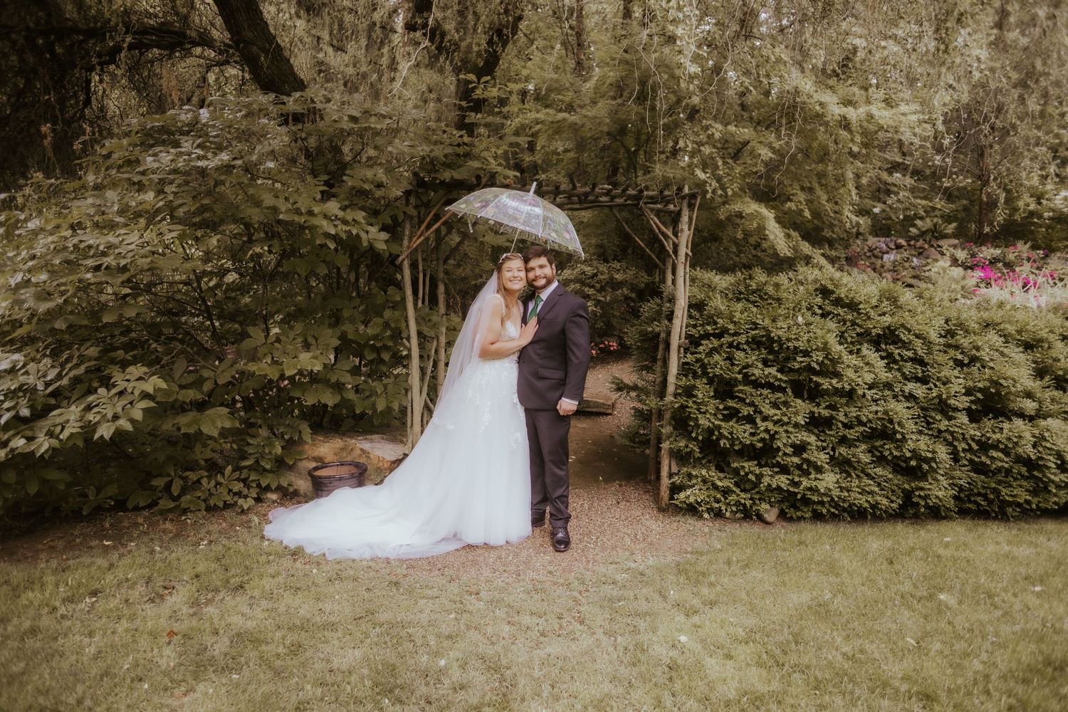 Bride and groom share a romantic embrace under a clear umbrella in a lush garden setting on their wedding day.