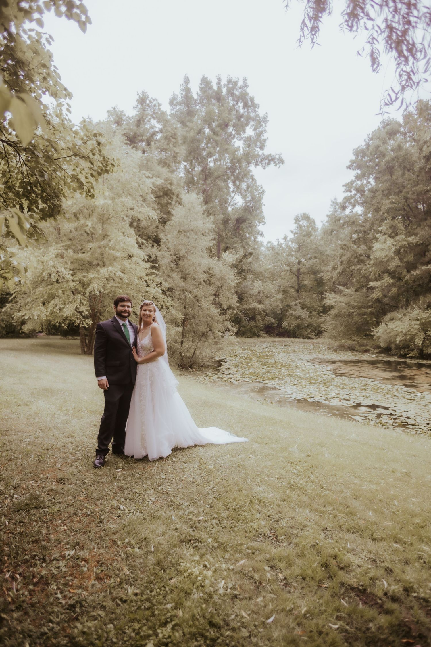 A bride and groom pose together on a lush green lawn beside a peaceful creek, surrounded by trees.