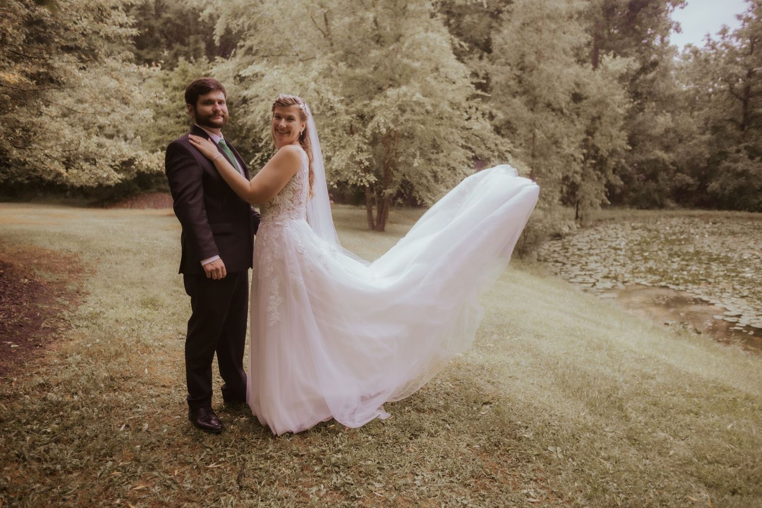 Bride in flowing white gown with veil blowing in wind stands with groom in dark suit outdoors on green lawn.