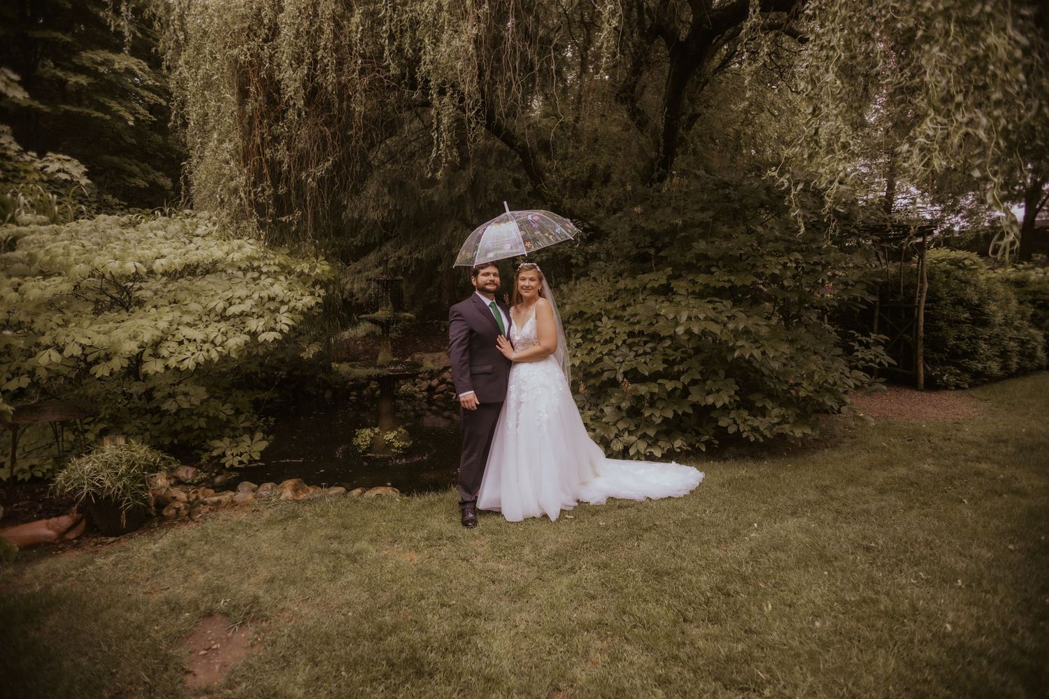 Bride and groom share an umbrella in lush garden on rainy wedding day, bride in white gown with train.