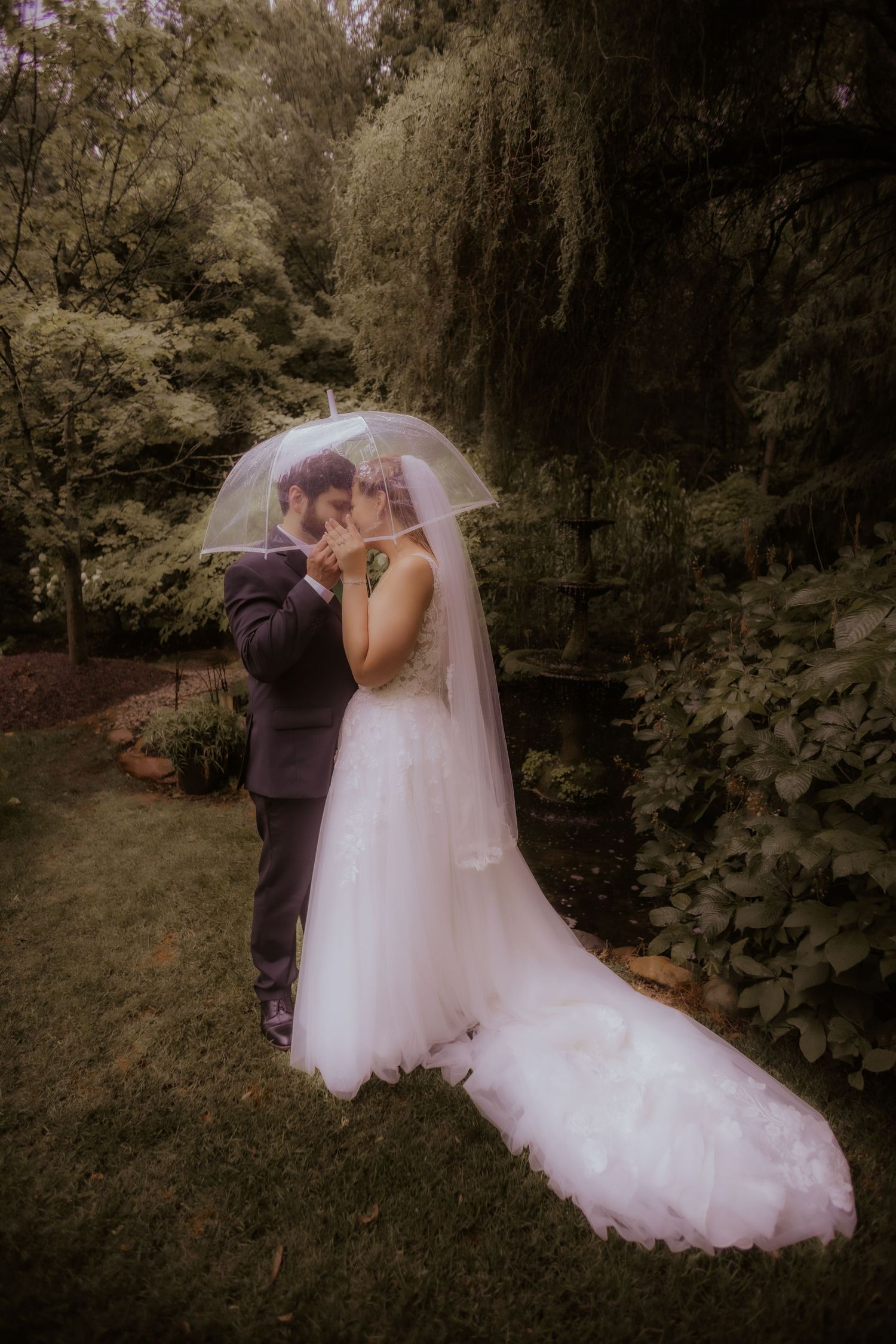 Bride and groom share a romantic kiss under a clear umbrella in a lush garden on their wedding day.