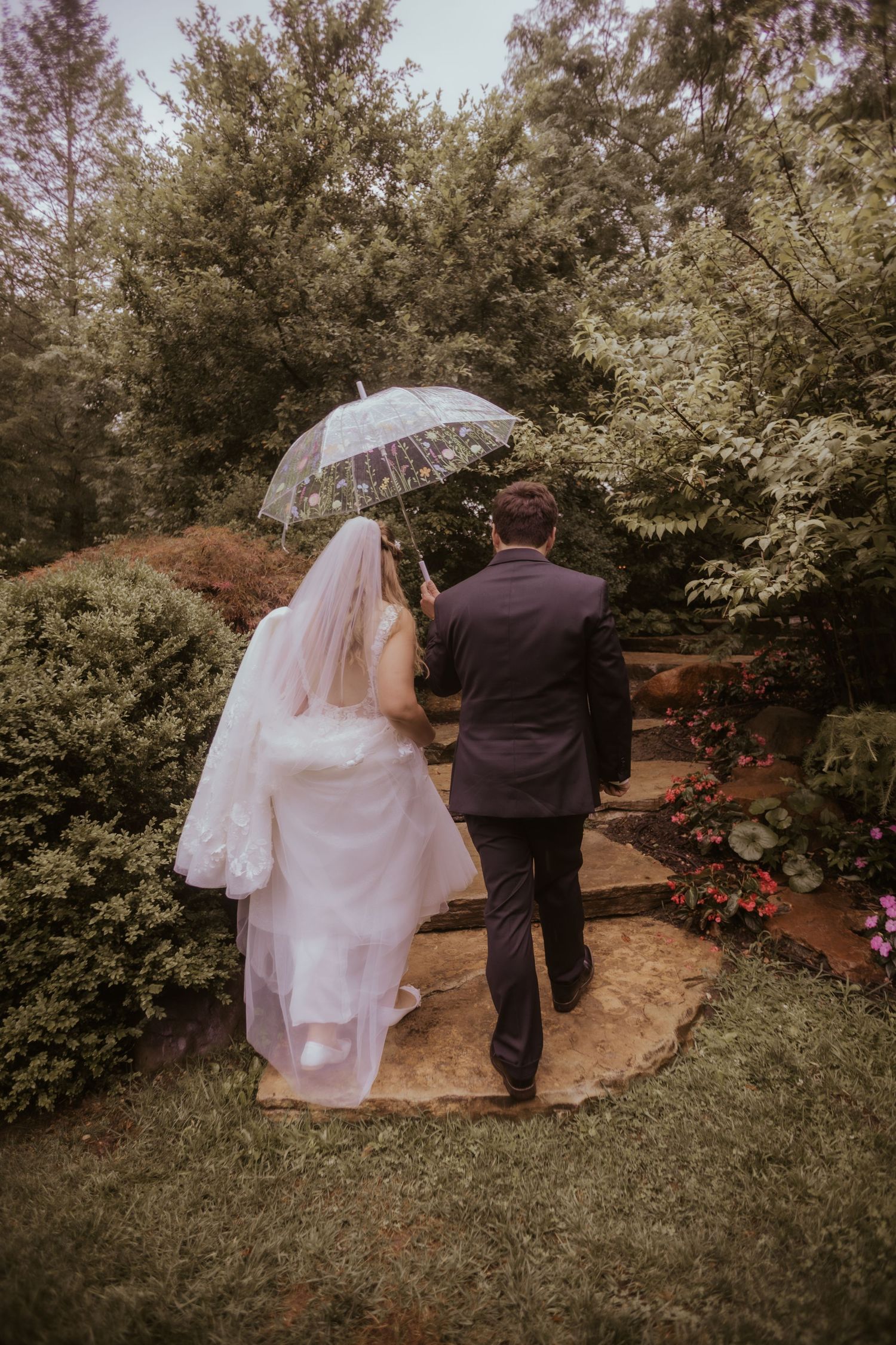 Bride and groom walking hand-in-hand through a lush garden in the rain, sharing a clear umbrella on their wedding day.