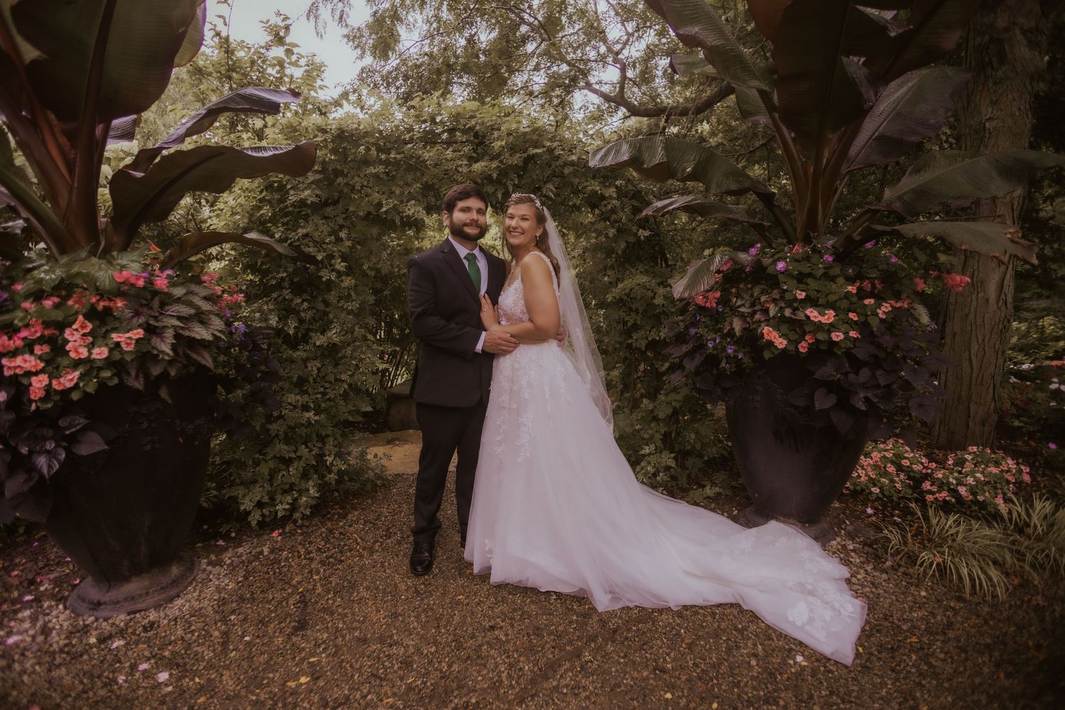 Bride and groom posing in a lush garden surrounded by pink flowers and greenery on their wedding day.